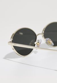 Gafas de sol redondas con lentes oscuros, montura de metal dorado y almohadillas nasales ajustables. Presenta varillas finas y un logo grabado en el lateral.
