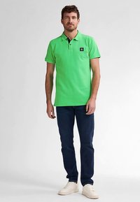 Zielona koszulka polo z krótkim rękawem, kołnierzyk na dwa guziki, z naszywką z logo na piersi, noszona z ciemnymi dżinsami i białymi trampkami. Gładka faktura materiału.