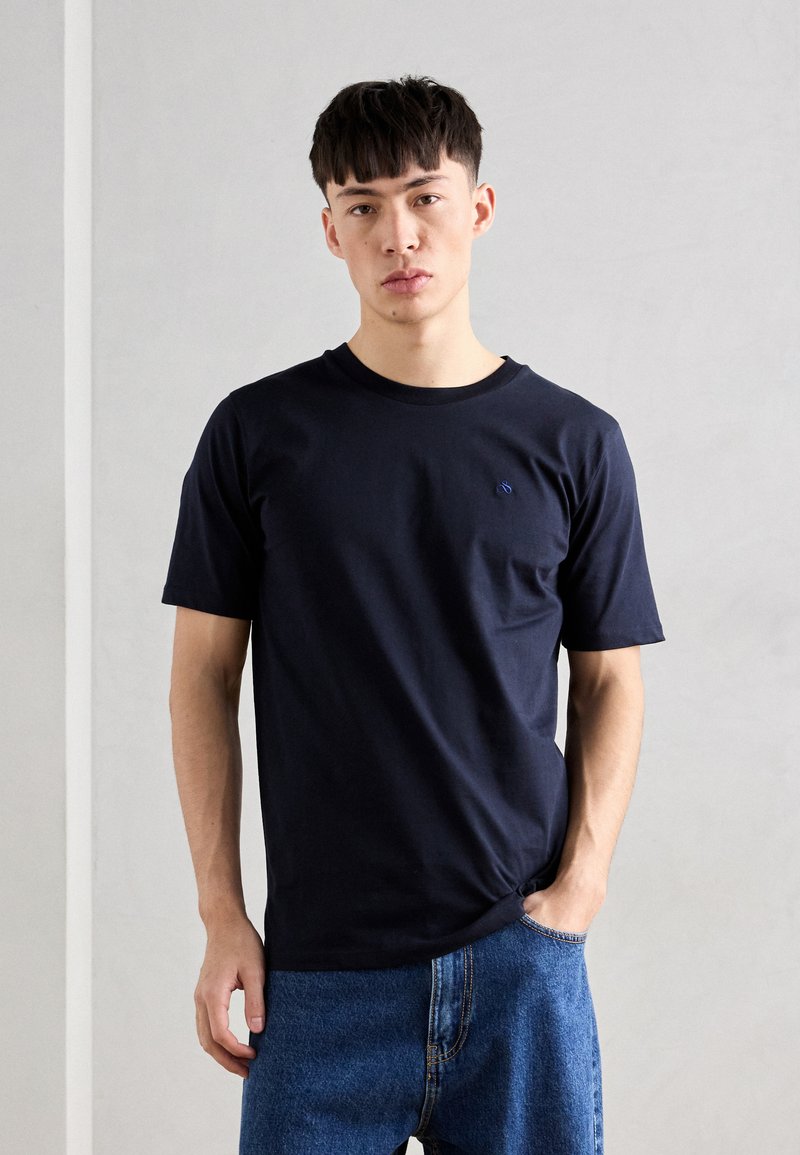 Tricou bleu marin cu mâneci scurte, realizat din bumbac, cu un guler rotund și un accent subtil al logo-ului pe piept, asortat cu blugi denim albaștri deschiși.