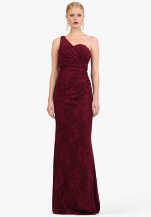 Frau trägt ein bodenlanges, einseitig schulterfreies, weinrotes Spitzenkleid, kombiniert mit kräftigem rotem Lippenstift und langen spiraligen Ohrringen.