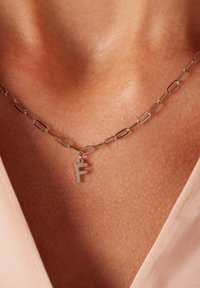 Zilveren ketting met verlengde schakels en een hangende metalen letter "F" hanger. De achtergrond heeft een huidskleur en zacht materiaal.
