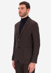 Uomo che indossa una giacca e un pantalone a coste marrone con un dolcevita nero, in piedi di fronte a uno sfondo bianco semplice.