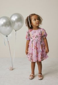 Blumenkleid in Hellrosa, mit Puffärmeln, gerafftem Taillenbereich und fließendem Saum. Kombiniert mit glitzernden pinken Schuhen und silbernen Ballons.