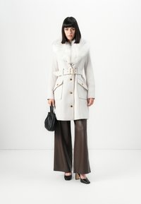 Vrouw in een witte gewatteerde jas met bontkraag, bruine wijde broek, zwarte hakken en een kleine zwarte handtas voor een eenvoudige achtergrond.
