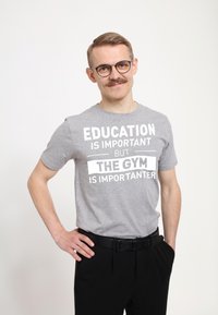Camiseta de algodón gris con texto blanco en negrita: "LA EDUCACIÓN ES IMPORTANTE, PERO EL GIMNASIO ES MÁS IMPORTANTE." Diseño de manga corta y cuello redondo.