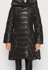 Giacca trapuntata nera con finitura lucida, caratterizzata da un motivo a rombi trapuntati, cappuccio oversize e maniche larghe. Rifinita con cuciture orizzontali.