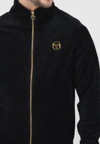 Schwarze Fleecejacke mit hohem Kragen, goldenem Reißverschluss und kleinem gesticktem Logo auf der linken Brust. Weicher Stoff und bequemer Sitz.