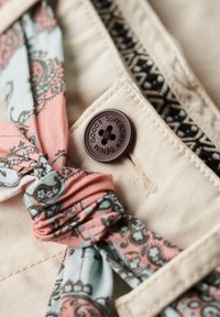 Beige stof met een bruine knop met het label "SUPERDRY DENIM." Een lichtgewicht stoffen sjaal met een patroon in roze, blauw en zwart is er dichtbij geknoopt.