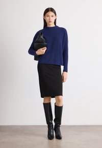 Blauer, kuscheliger Pullover, schwarzer, taillierter Midirock, kniehohe schwarze Stiefel und eine schwarze Clutch. Minimalistisches Design mit sanften Texturen.