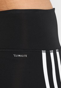 Černé sportovní legíny se širokým pasem. Na bocích mají logo Climalite a tři bílé pruhy. Hladká textura.