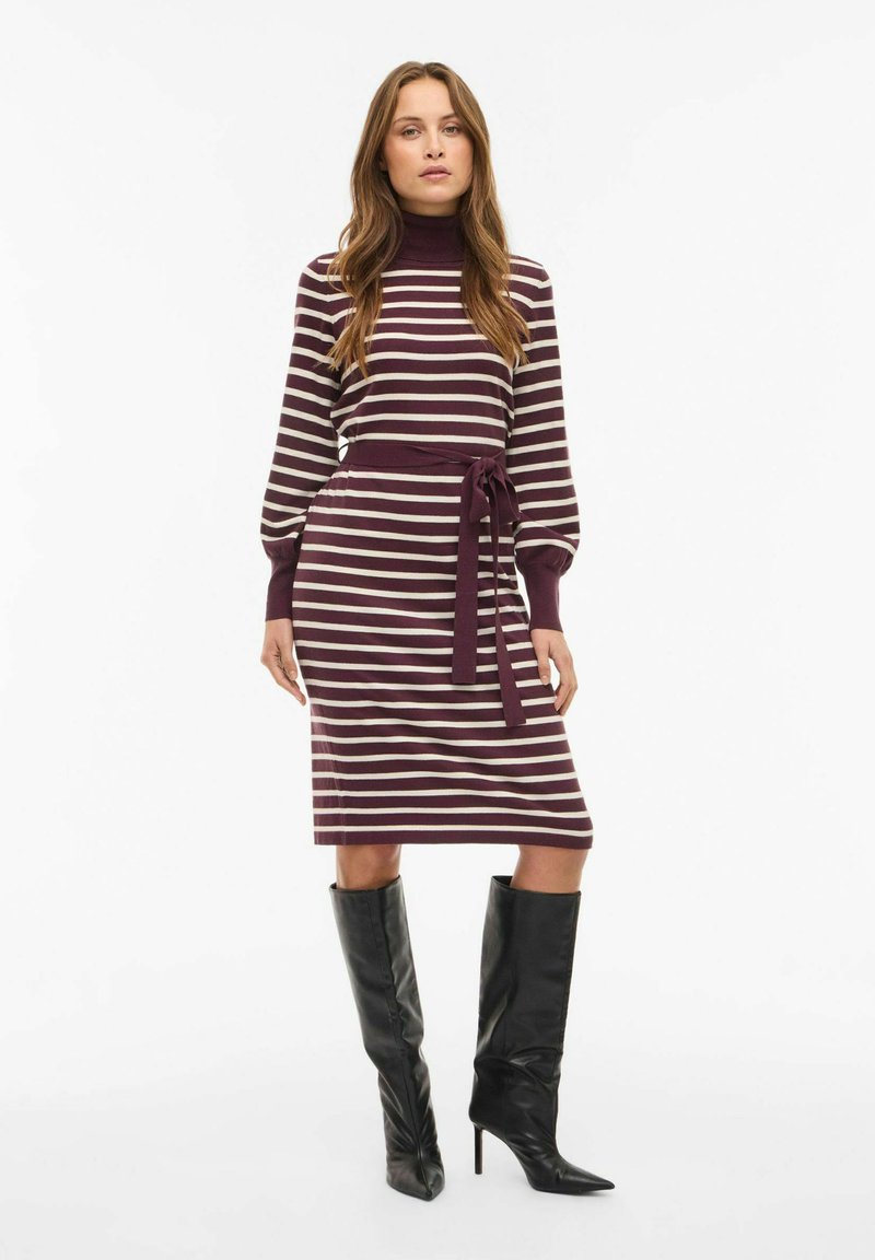 Kootud kleit burgundi ja kreemivärvi horisontaalsete triipudega, kõrge kaelusega, kitsas talje ja vastav nööri vöö; koos põlvikute mustade saapatega.