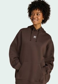 Sudadera de gran tamaño en color marrón con un bolsillo delantero y capucha con cordón. Presenta un logo de adidas en blanco en el pecho. Tejido suave.