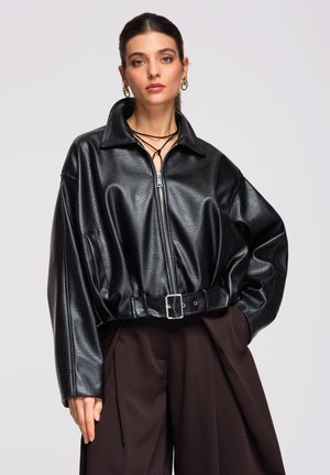 Femme aux cheveux tirés en arrière portant une grande veste en cuir noire zippée avec ceinture et un pantalon large foncé, devant un fond gris uni.