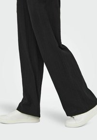 Pantalon large noir en tissu texturé, présenté avec des baskets blanches. Le pantalon a un motif rayé vertical subtil.