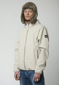 Veste imperméable beige avec capuche, dotée d'un zip frontal complet, de deux poches latérales avec bord coloré et de poignets élastiques. Bonnet doublé en fourrure inclus.