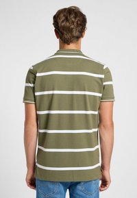 Polo de color verde oliva con rayas horizontales blancas. Mangas cortas, cuello acanalado y aberturas en los laterales del dobladillo. Textura suave.