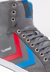 Grå høj-top sneakers med røde og blå detaljer, der har metaløjer, en tekstureret stofoverdel og et logotab på tungen.