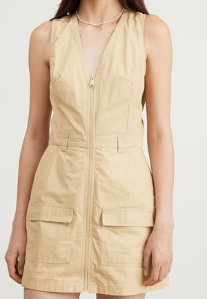Robe sans manches couleur sable avec un décolleté en V profond, une fermeture éclair à l'avant, deux poches plaquées à l'avant et une taille cintrée. Confectionnée dans un tissu léger.