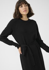 Schwarzes langärmliges Kleid aus weichem Stoff, mit einem runden Halsausschnitt und einem gebundenen Gürtel in der Taille. Geschmeidige Textur mit taillierten Manschetten.