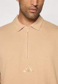Beige polo shirt met korte mouwen en een gestructureerd, wafelpatroon, voorzien van een rits aan de voorkant en een klein, geborduurd logo op de borst.