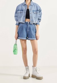 Veste en denim cropped, bleu clair, avec des bords décolorés et effilochés ; accompagnée de shorts en denim taille haute ; tenant un sac à main vert ; portant des bottes à plateforme blanches.