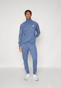 Blå joggedress i glatt stoff, med en jakke med glidelås og avsmalnende bukser med hvite aksenter langs ermene og sidene.