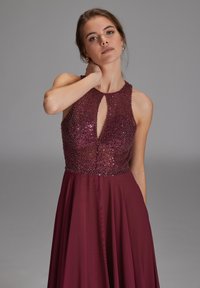 Robe de soirée bordeaux avec un corsage sans manches orné de perles, présentant un décolleté en trou de serrure et une jupe fluide en transparence.