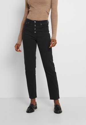 Osoba ubrana w czarne dżinsy z wysokim stanem z widocznymi guzikami i postrzępionymi krawędziami, w połączeniu z beżowym prążkowanym topem z długim rękawem i czarnymi mokasynami.