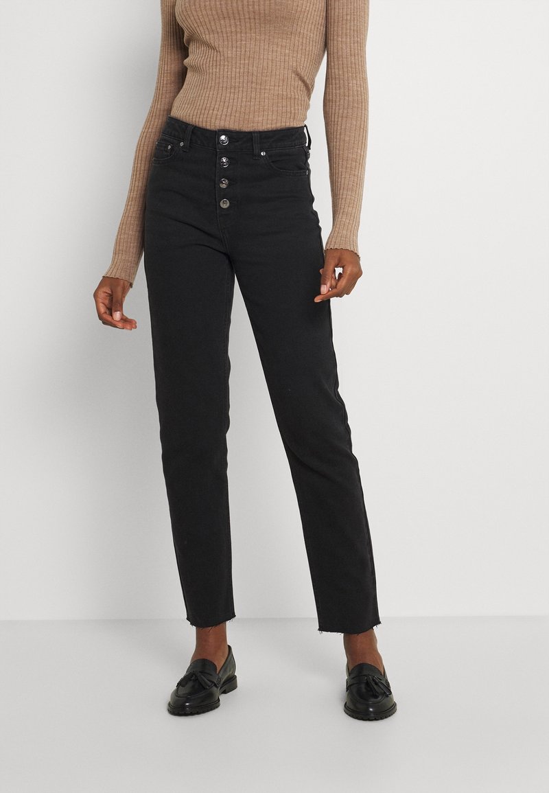 Personne portant un jean noir taille haute avec des boutons apparents et des ourlets effilochés, associé à un haut beige à manches longues côtelé et des mocassins noirs.