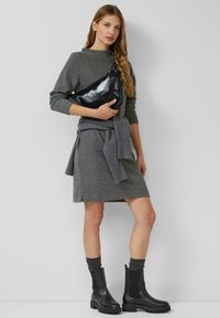 Grijze trui en jurk, gecombineerd met een zwarte glanzende schoudertas. Zwarte enkellaarsjes met getextureerde zolen en grijze gebreide sokken maken de outfit compleet.