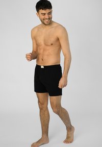 Černé bavlněné boxerky s knoflíkovým zapínáním, elastickým pasem a štítkem na pasu, vystavené na muži, který stojí bos.