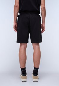 Black cotton shorts with an elastic waistband, side pockets, and a relaxed fit. Worn with beige sneakers and black socks.