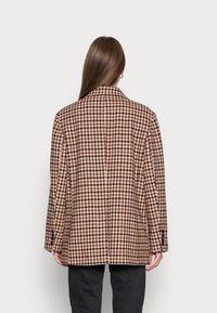 Koszula overshirt z mieszanki wełny w kratę z kołnierzem, wentylacją z tyłu i guzikami przy mankietach. Paleta kolorów obejmuje odcienie brązu, kremu i czerni.