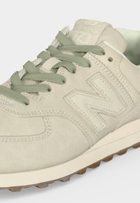 Baskets New Balance beige clair avec texture en daim, lacets en tissu verts, logo en relief sur le côté et bord de semelle en caoutchouc texturé visible.