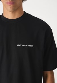 Camiseta de algodón negra con texto bordado en blanco "no desperdicies la cultura" en el lado izquierdo del pecho. Cuello redondo y mangas cortas.