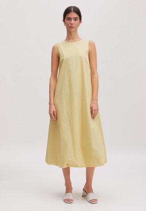 Vrouw draagt mouwloze, loszittende, lichtgele midijurk met ronde halslijn en witte open teen sandalen met hak, staand tegen een effen achtergrond.