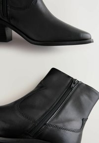 Botines de cuero negro con punta afilada, cierre de cremallera y acabado texturizado. Cuentan con un tacón apilado y costuras decorativas.