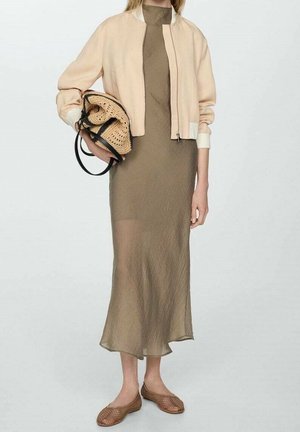 Femme portant une veste courte beige sur une robe midi olive, tenant un sac tissé et portant des ballerines marron tissées sur un fond uni.