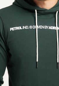 Felpa verde in tessuto morbido con logo stampato bianco, cordini regolabili beige e punte metalliche sui cordini.