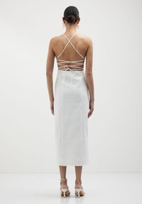 Abito midi bianco con spalline incrociate sul retro, texture liscia e dettaglio annodato in vita. Combinato con sandali con tacco a blocco.