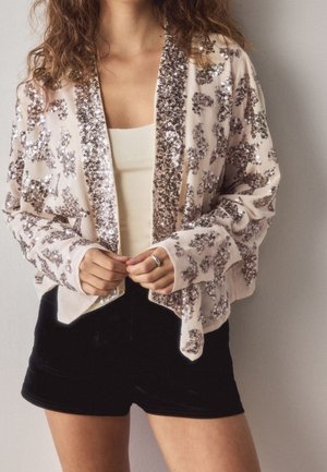 Femme portant une veste rose clair à motif pailleté par-dessus un haut blanc et un short noir, debout contre un mur uni.