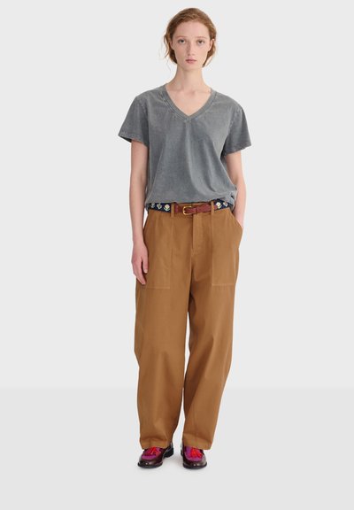 Jeune personne debout, les mains dans les poches, portant un t-shirt gris délavé à col en V, un pantalon large marron, une ceinture florale et des chaussures violettes avec des pompons roses.