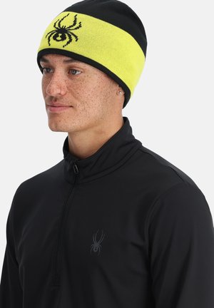 Berretto lavorato a maglia giallo e nero con logo di ragno, indossato con un top nero a zip quarti. Texture liscia con vestibilità aderente.