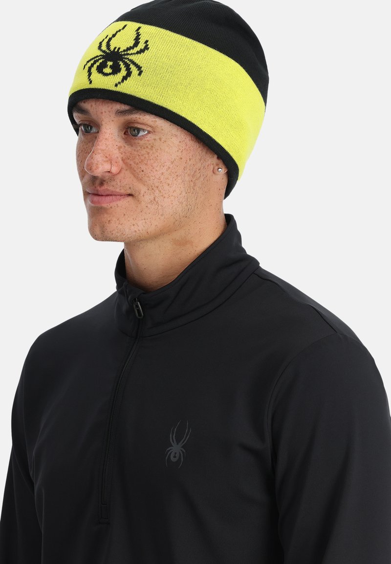 Berretto lavorato a maglia giallo e nero con logo di ragno, indossato con un top nero a zip quarti. Texture liscia con vestibilità aderente.