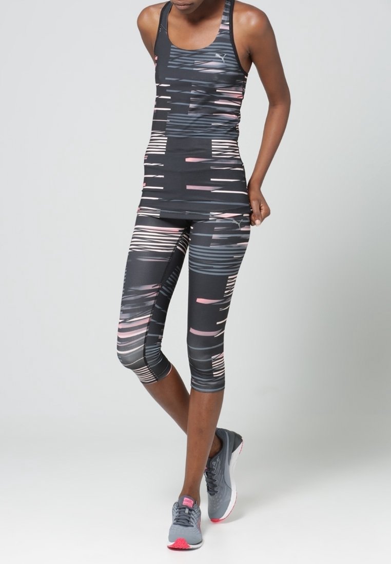Femme portant un débardeur de sport à motifs noirs, gris et roses et des leggings capri avec des chaussures de course grises, debout devant un fond neutre.