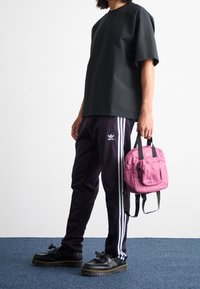 Osoba v černých kalhotách Adidas s bílými pruhy, černém tričku, černých botách, držící malý růžový batoh za držadla.