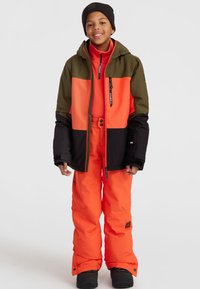 Veste isolante orange vif et olive avec des accents noirs, assortie à un pantalon orange et des bottes noires. Comprend des zips et des pressions.
