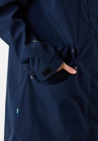 Navy waterproof jacket featuring a front pocket with snap closure, adjustable cuff with Velcro, and a small Swedish flag accent near the hem.