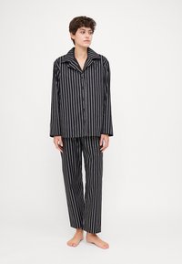 Musta-valkoinen raidallinen pyjamapuku, joka sisältää napitettavan takin leveällä kauluksella ja siihen sopivat suorat housut, valmistettu pehmeästä kankaasta.