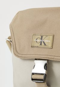 Bežová látková messenger taška s textúrovaným povrchom, striebornými doplnkami a nášivkou s logom "Calvin Klein Jeans" v minimalistickom dizajne.